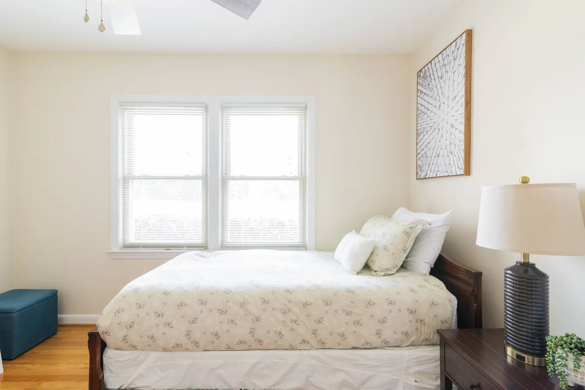 A tranquil bedroom featuring a bed with floral bedding, a cozy accent chair, a nightstand with a lamp, and artwork. Sunlight filters in through the windows, creating a warm and welcoming atmosphere ideal for relaxation and unwinding.