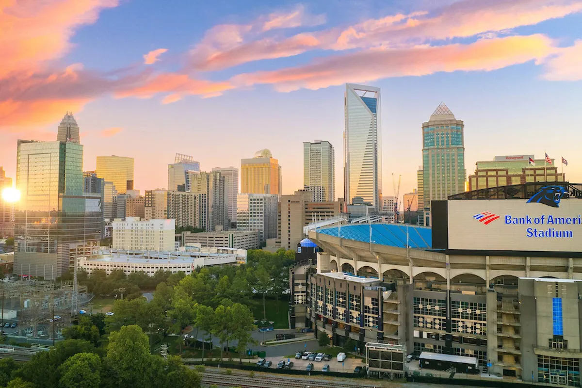 The vibrant Charlotte skyline glows at sunset, with Bank of America Stadium in view. The scene is energetic, hinting at the city's excitement, sports events, and urban outings just a short drive from Peachtree Hideaway.