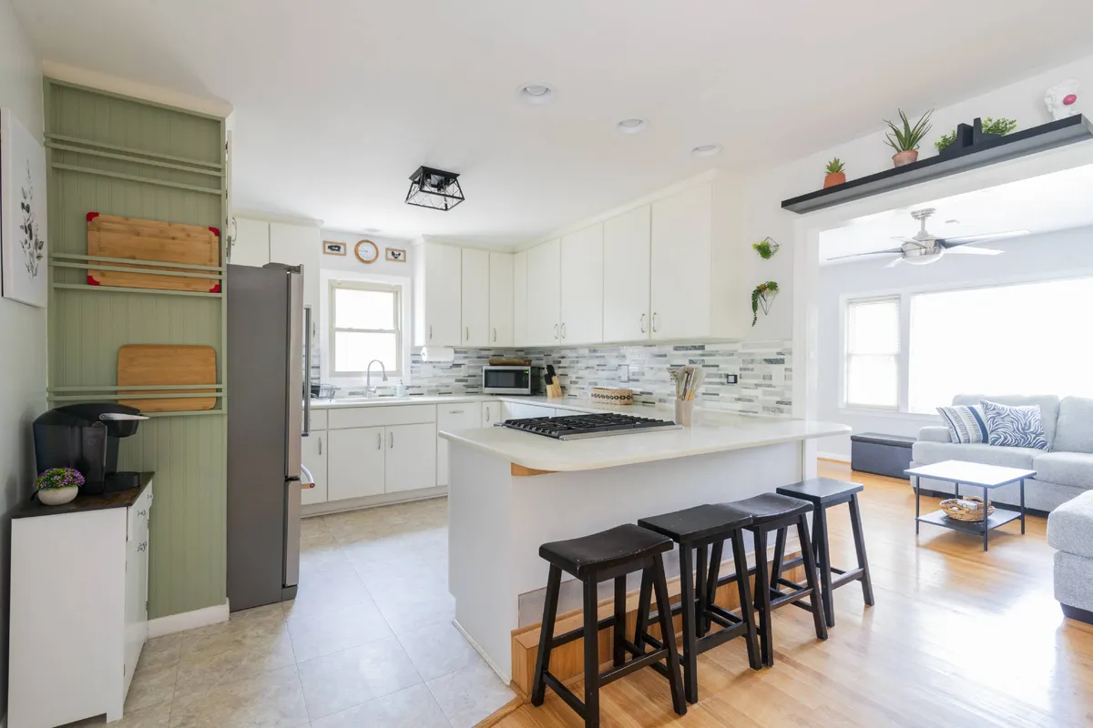 This image features a kitchen island with stools, modern kitchen appliances, and generous natural light streaming in through the windows. The space is open and inviting, with clear areas for both cooking and gathering.