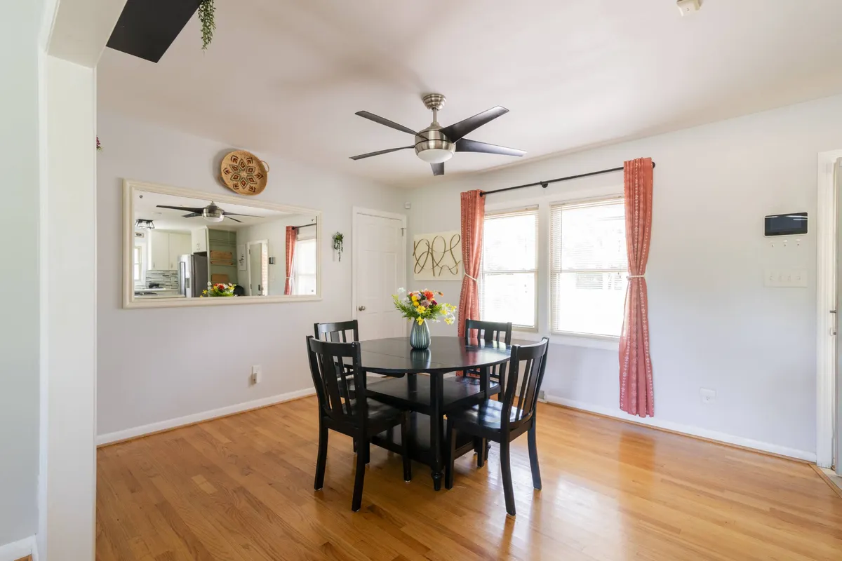 An inviting dining nook with a wooden table adorned with flowers in a vase, a mirror reflecting the cozy interior, and sheer curtains letting in natural light. The intimate arrangement is perfect for shared meals or casual conversations.