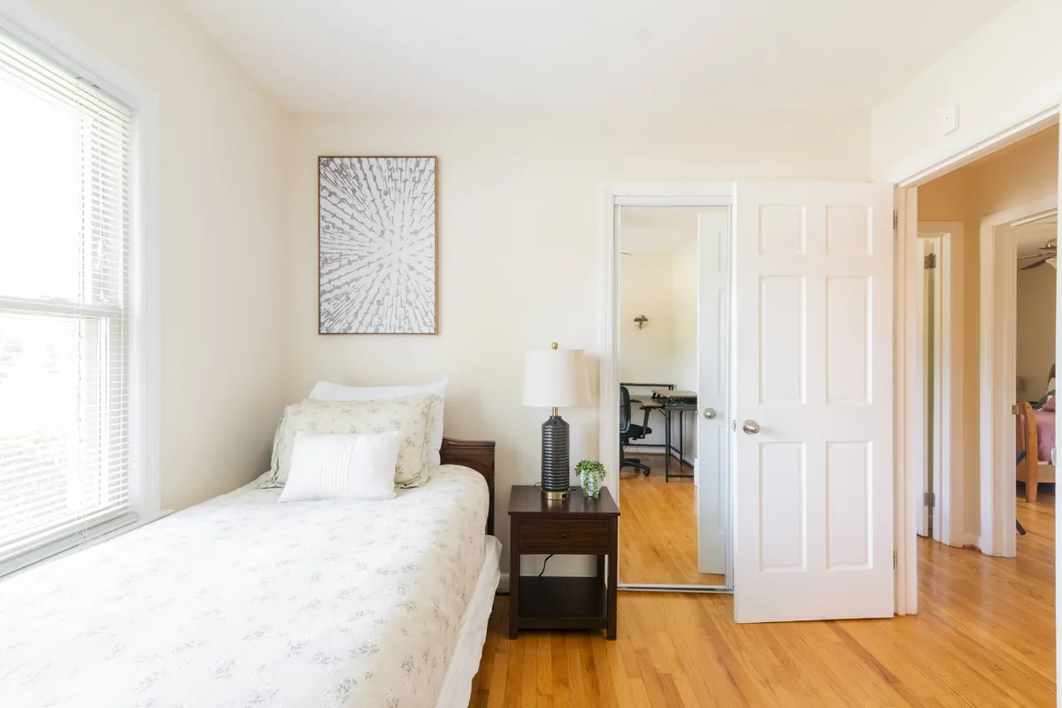 This serene bedroom offers a bed with floral bedding, a nightstand with a lamp and plant, wall art, and an open door leading to another room—suggesting spaciousness and easy flow through the home.