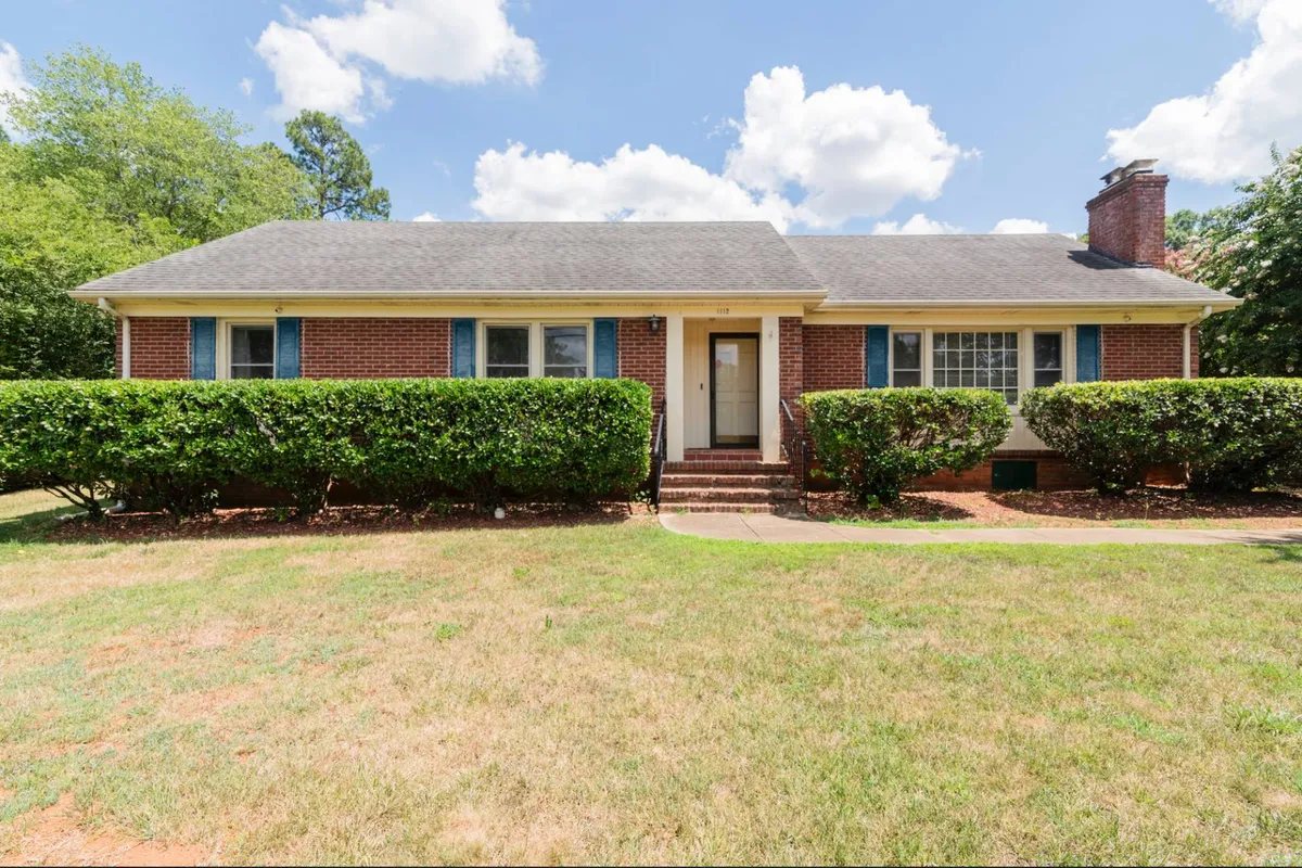 This image depicts the exterior of Peachtree Hideaway, featuring its brick facade, blue shutters, and neatly trimmed bushes. The spacious front yard and inviting porch hint at the property's welcoming and family-friendly environment. The scene suggests spaces where children and pets might play together and families can gather outdoors.