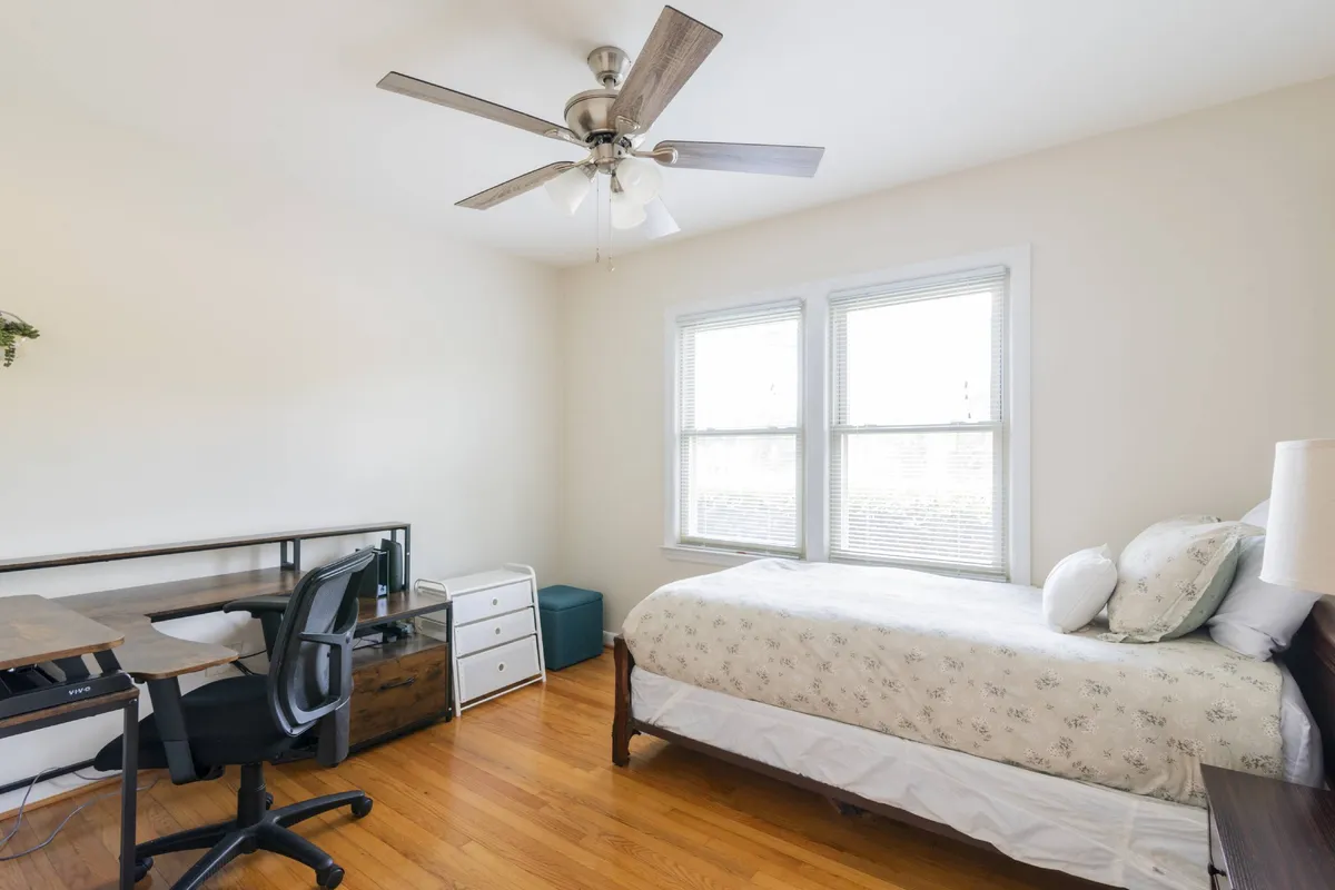 A serene bedroom with floral bedding, a desk and chair, and large windows that flood the space with morning light. The room is styled for both relaxation and productivity, with a cozy reading nook by the bed and an inviting workspace near the window.