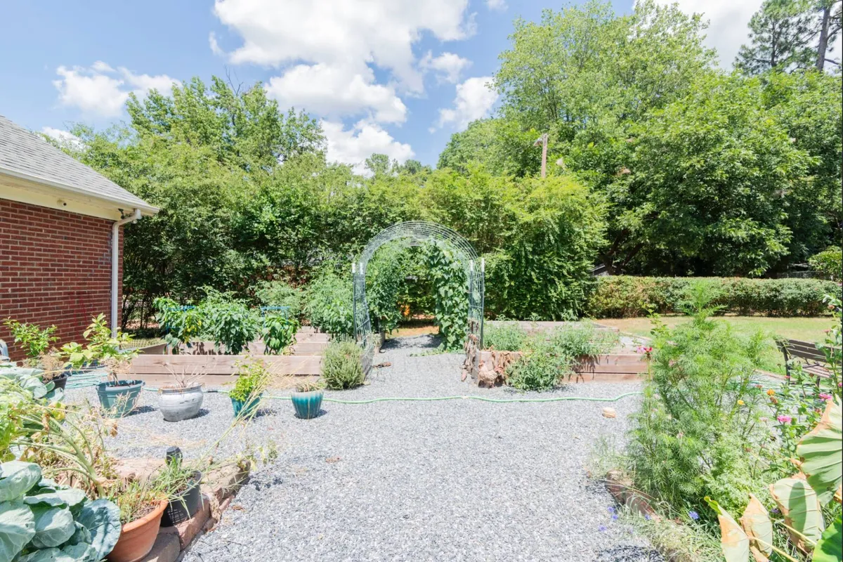 A peaceful garden scene with a charming archway leads to vegetable beds brimming with a variety of plants. The area is clearly meant for both relaxation and hands-on gardening, with plenty of natural beauty and a sense of tranquility.