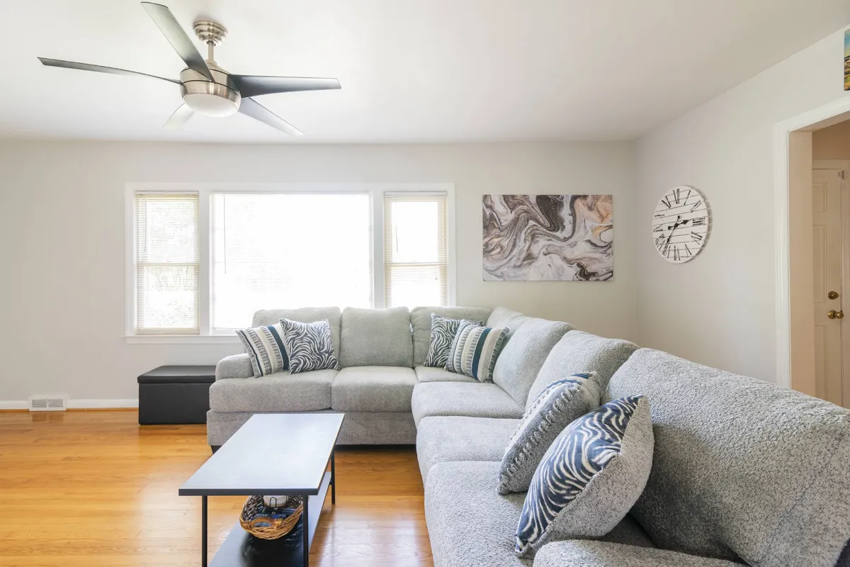 A bright and airy living room with a large sectional sofa positioned near multiple windows, a coffee table, wall art, and a ceiling fan. The natural light and open layout convey a sense of flexibility and comfort, ideal for both socializing and unwinding.